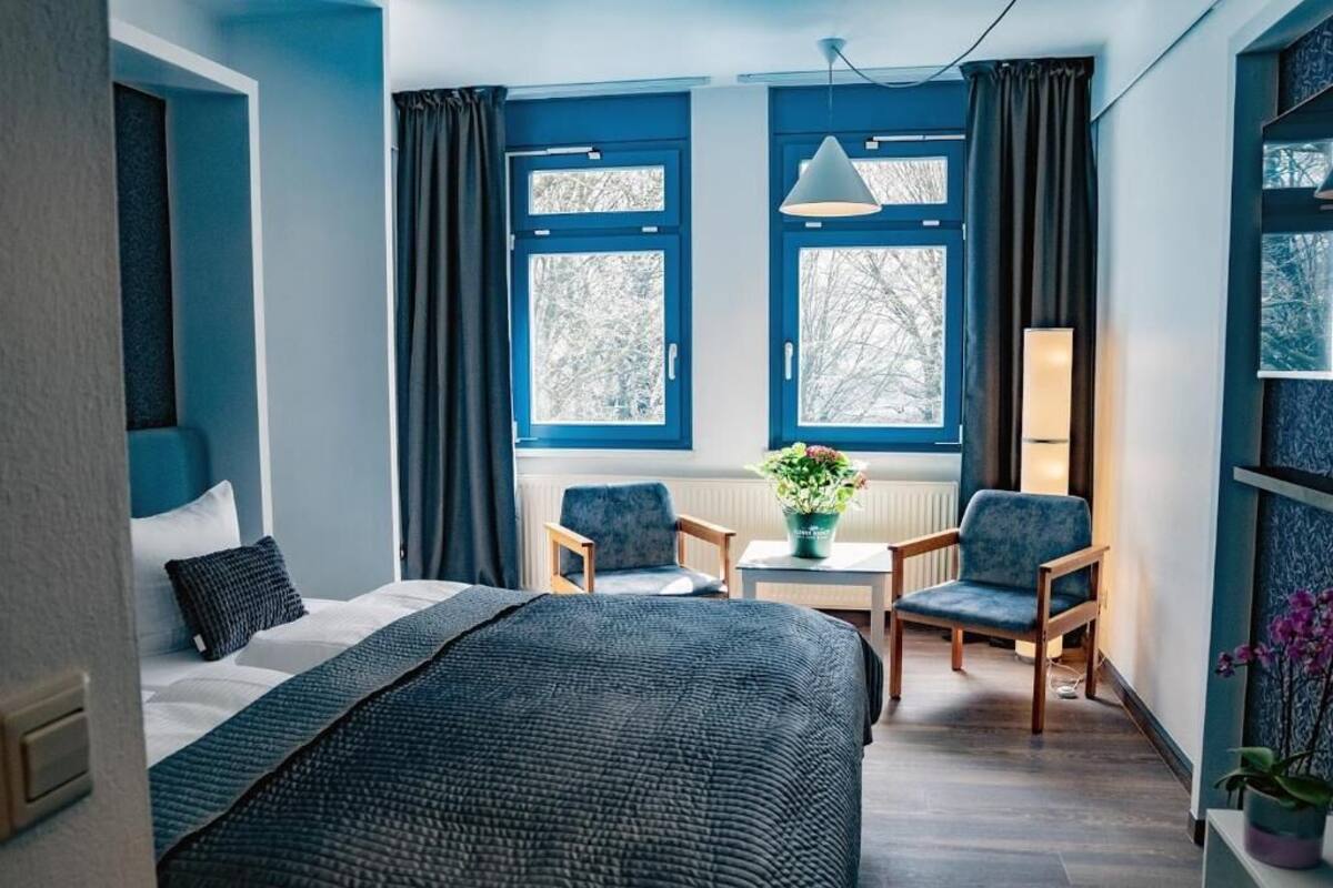 Een slaapkamer met een bed, twee stoelen, een tafel, een kamerplant en een raam met gordijnen.