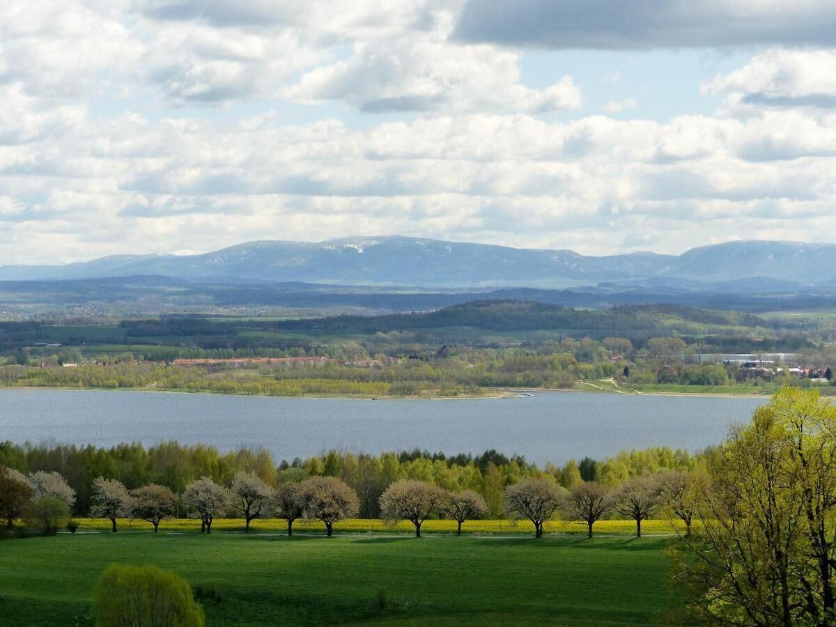 Un lago rodeado de campos verdes y árboles, con montañas distantes y un cielo nublado.