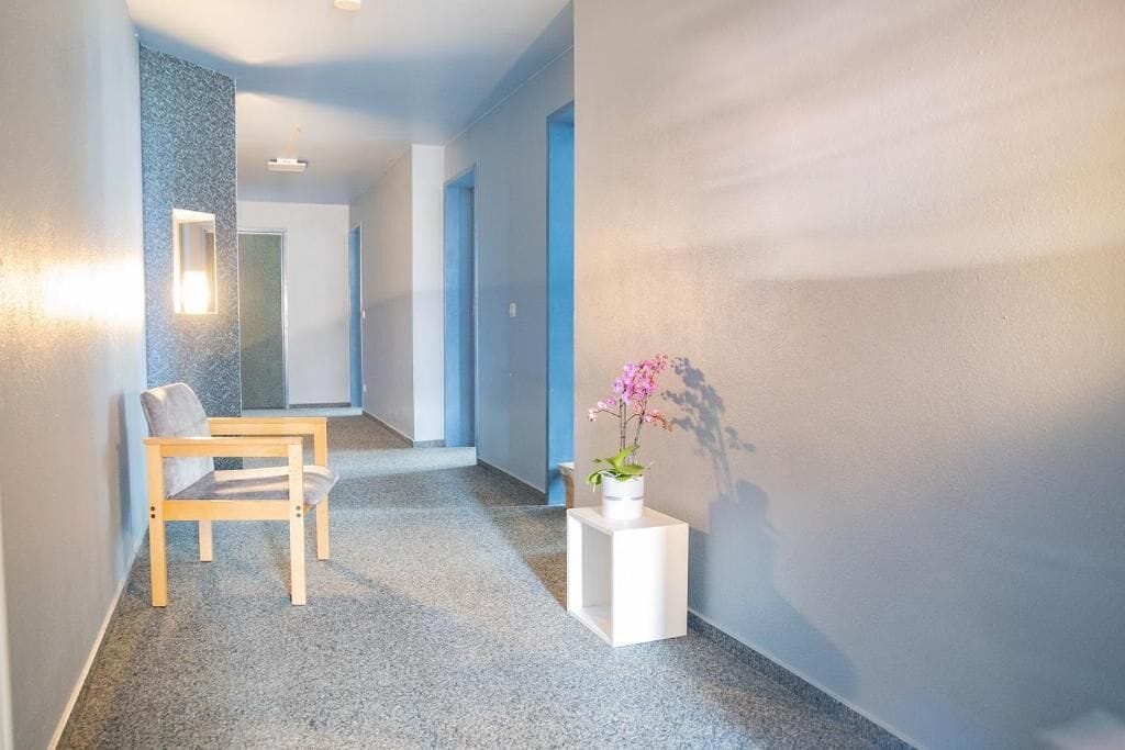 Un pasillo con una silla de madera, una mesita auxiliar blanca con planta en maceta y un espejo en la pared.