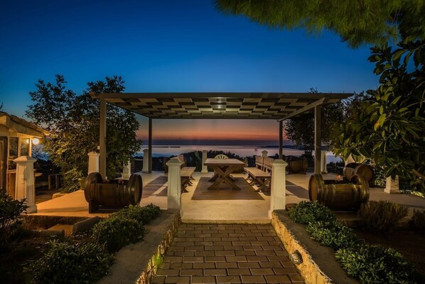 An outdoor patio with a pergola, seating, and barrels, overlooking a scenic view.