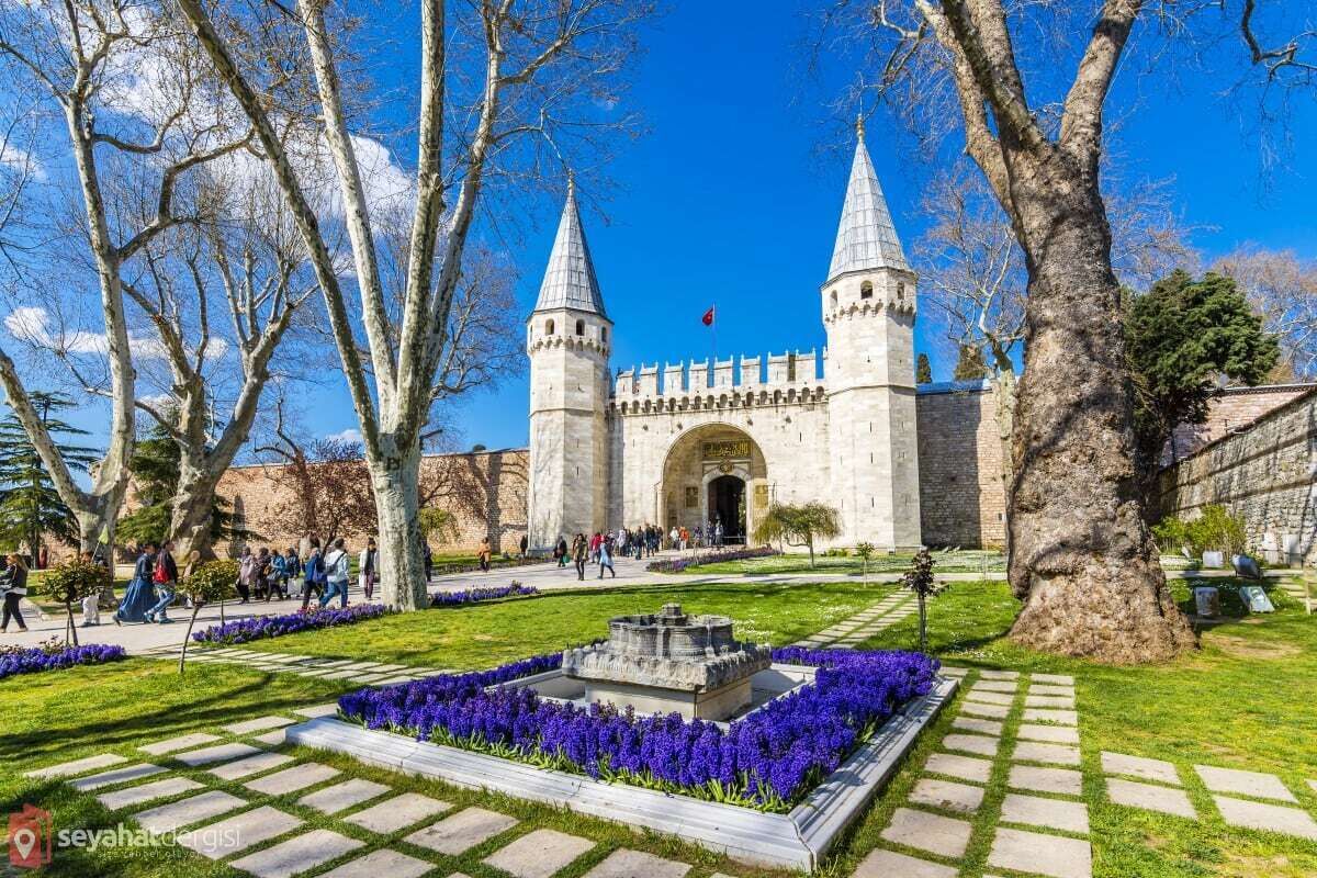 A historic building with two towers, surrounded by a well-maintained garden with purple flowers and a central fountain.