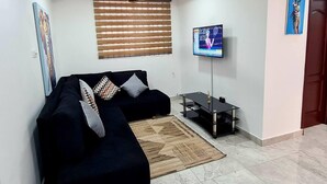 A living room with a dark sofa, a flat-screen TV, a ceiling fan, and a window with blinds. - Spacious, Quiet & Cozy- Close to Highway-parking (Adenta)