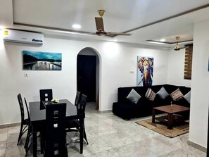 A modern living room with a dining area, featuring a black sofa, a coffee table, and a dining table with chairs. There are ceiling fans, a painting of an elephant, and a painting of a pier. - Spacious, Quiet & Cozy- Close to Highway-parking (Adenta)