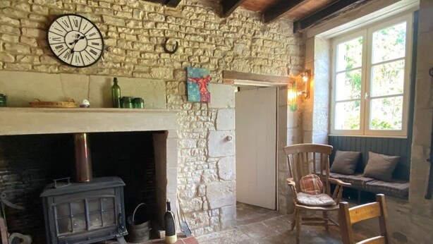 A rustic room with a stone fireplace, a wooden chair, and a window with a view of greenery. - Beautiful country house with private garden and heated swimming pool (Dampierre-sur-Boutonne)