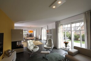 A modern living room with a sofa, two armchairs, a coffee table, and a fireplace. There is a dining area in the background with a table and chairs.