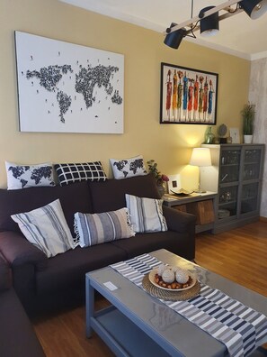Een woonkamer met een donkerbruine bank, een wereldkaart kunstwerk, een kleurrijk abstract schilderij, een lamp, een kast met glazen deuren, en een gestreept tafelkleed.