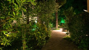 Un camí de jardí nocturn amb paviment de maó, envoltat de vegetació densa i il·luminat per una llum verda ambiental.