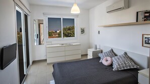A modern bedroom with a bed, bedside tables, a dresser, a TV, and a window with a view. - Protaras holiday villa - rentprivatevillas 145 (Protaras)