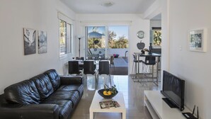 A modern living room with a black leather sofa, a coffee table with a bowl of fruit, a flat-screen TV, and a dining area with chairs and a table. - Protaras holiday villa - rentprivatevillas 145 (Protaras)