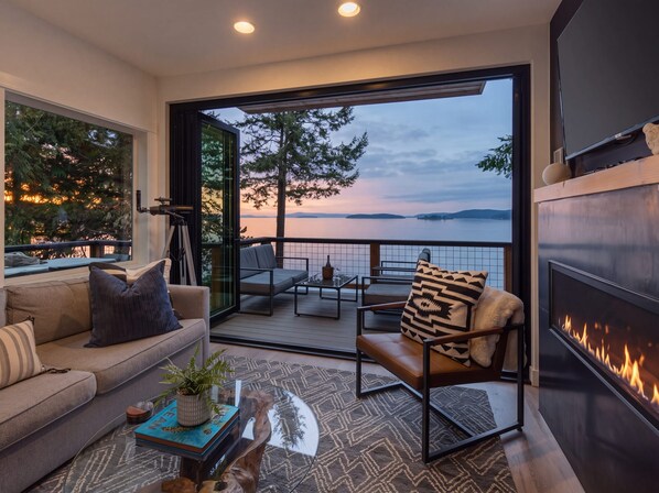 A modern living room with a sofa, coffee table, and a fireplace. There is a balcony with a view of the water and trees.
