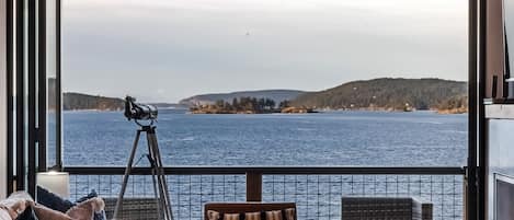 A balcony with a telescope, comfortable seating, and a view of a large body of water and distant land.