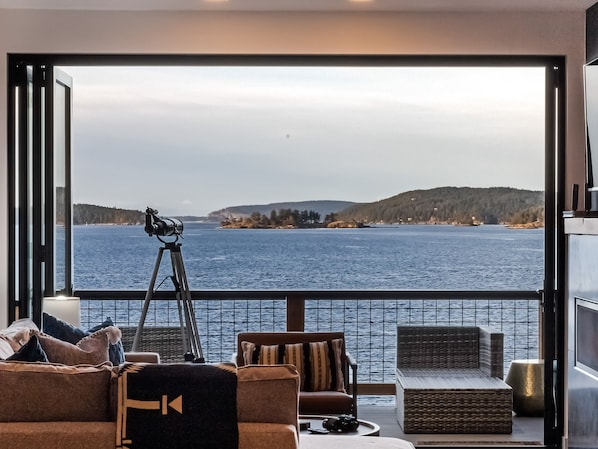 A balcony with a telescope, comfortable seating, and a view of a large body of water and distant land. - Waterfront Home! Hot Tub! (HOS) (Friday Harbor)