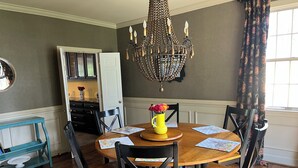 A dining room with a round wooden table, black chairs, a chandelier, and a window with patterned curtains.