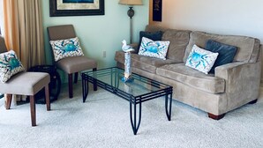 A living room with a beige sofa, a glass coffee table, a framed painting, and a lamp. - 3 Bedroom Direct Ocean Front Condo in Silver Moon with Outdoor Pool! (Ocean City)