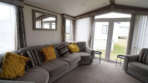 A living room with a grey sectional sofa, a small round table, and a window with curtains. - Pets Stay Free 8 Berth lux van Heacham Beach (King's Lynn)