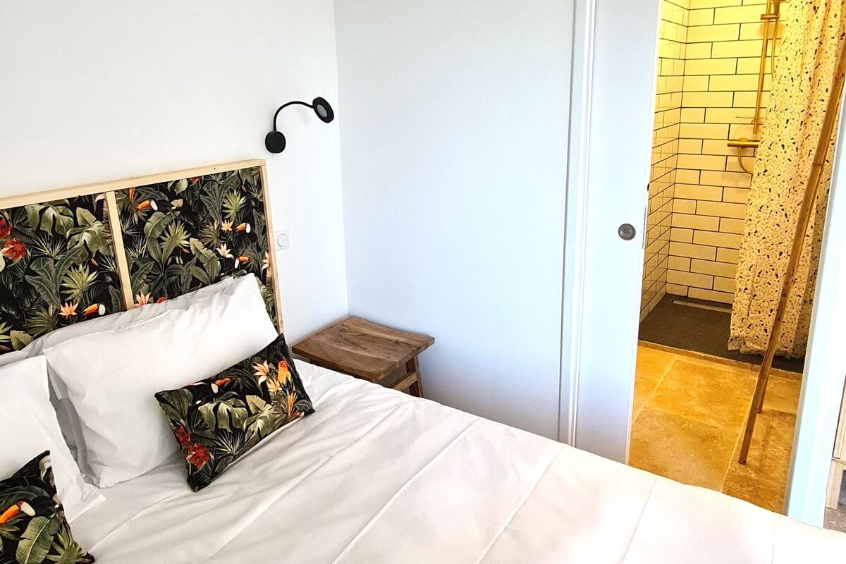 A neatly made bed with white linens and a floral-patterned pillow, a small wooden nightstand, and a wall-mounted light fixture.