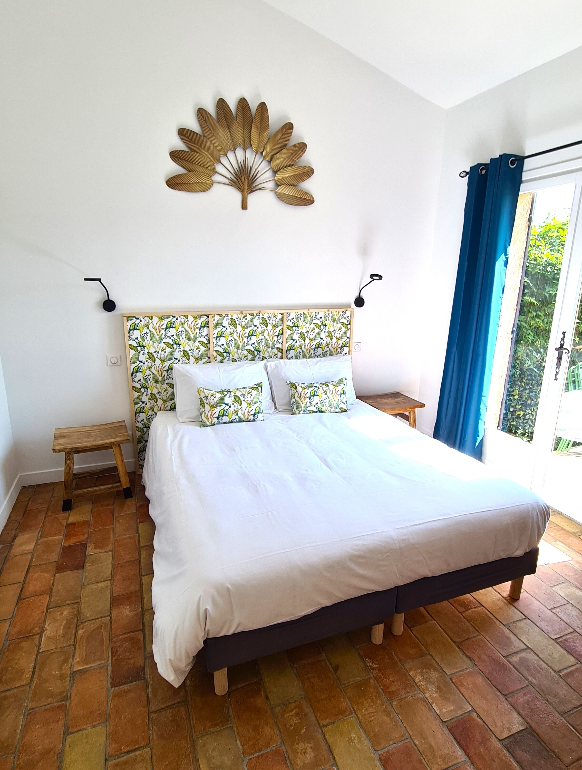 A bedroom with a bed, a wooden headboard with a floral pattern, a wall-mounted decorative piece, a small wooden stool, and a view of greenery through a window with blue curtains.