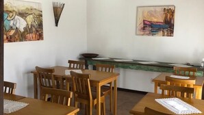 A dining area with wooden tables and chairs, a kitchen counter, and two paintings on the walls. - Pousada Casa da Aroeira (Búzios)