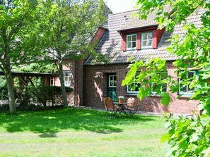 Une maison en briques avec un toit en pente, un petit porche avec une table et des chaises, et un jardin avec de la pelouse et des arbres.