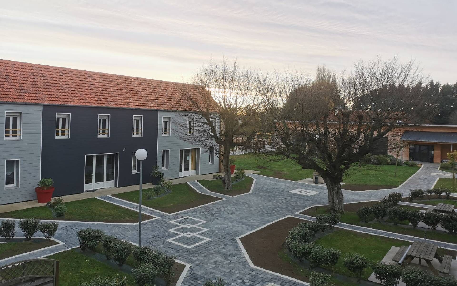 A modern building with a red-tiled roof, a paved courtyard with geometric patterns, and a well-maintained lawn.