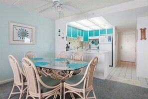 Un coin repas avec une table en verre et des chaises en osier, une cuisine avec des meubles de couleur turquoise et un ventilateur de plafond.