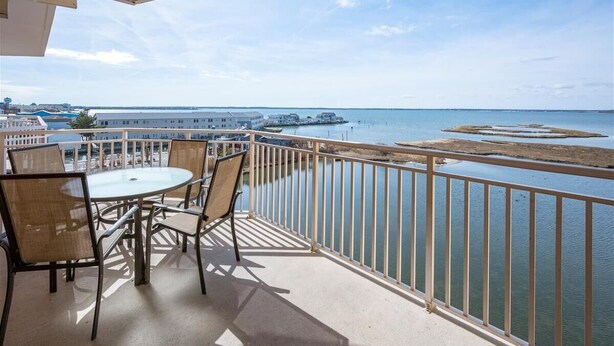 Un balcon avec une table et des chaises donnant sur le bord de l’eau, avec des bâtiments et une jetée.