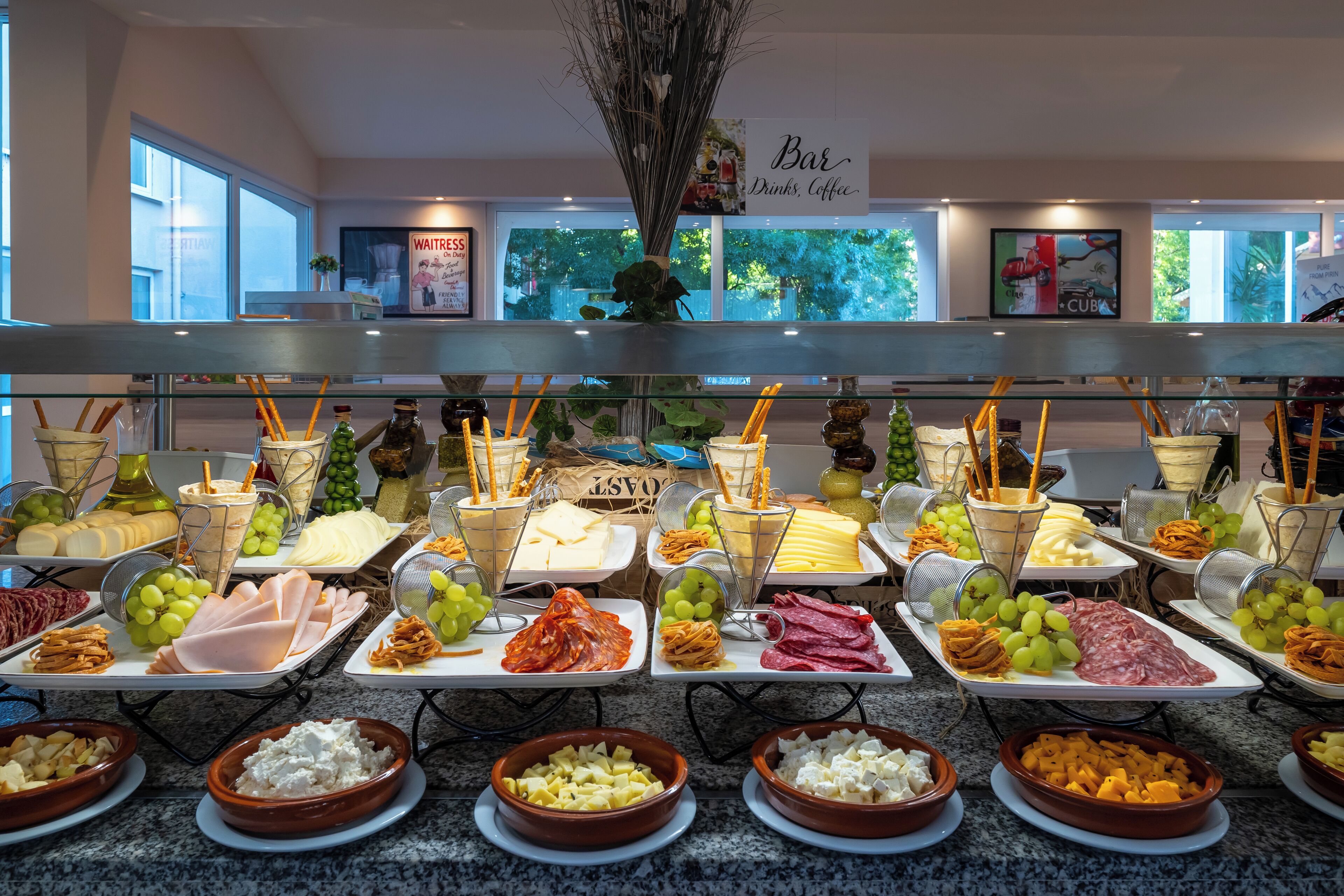 Un bufé con diversos alimentos y bebidas, incluyendo queso, carnes y frutas, exhibidos sobre un mostrador con un letrero que dice 'Bar Drinks, Coffee'.