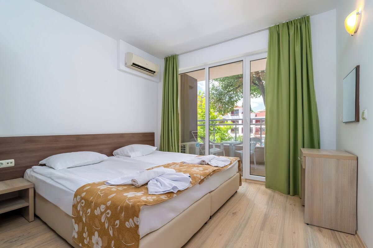 A hotel room with a bed, a wooden nightstand, a small wooden cabinet, and a sliding glass door leading to an outdoor area.