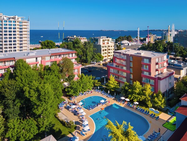 Ett semesterområde med en swimmingpool omgiven av solstolar och parasoller, beläget intill en hög byggnad och en vattenyta.