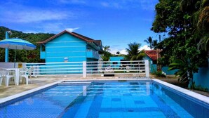 A swimming pool with a blue tiled deck, a white fence, and a blue building with a red roof in the background. - Chalés Céu e Mar (Ubatuba)