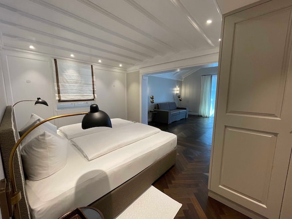 A modern hotel room with a bed, bedside lamps, a nightstand, a sofa, and a view of the adjacent room.