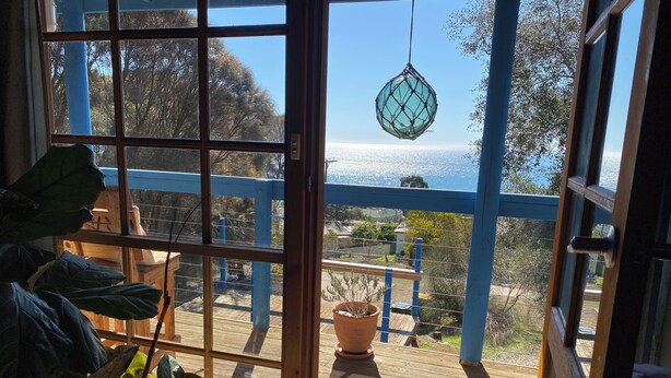 Uitzicht door een raam met blauwe lijst, een hangende macramé decoratie en een bloempot met plant op een houten terras.