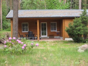 Una cabaña de madera con techo de tejas, un porche con mesa y sillas, y un pequeño jardín con flores rosadas.