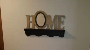 Un banc en bois avec un coussin appuyé contre un mur, et une pancarte en bois au-dessus, sur laquelle on peut lire le mot « MAISON ».