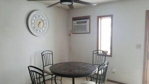 Un coin repas comprenant une table ronde et quatre chaises, un ventilateur de plafond, une horloge murale et une fenêtre avec des stores.