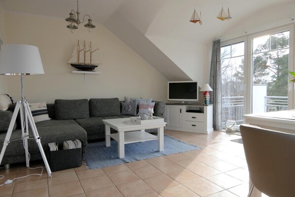 Ein modernes Wohnzimmer mit grauem Sofa, weißem Couchtisch und einem Fernseher auf einem Schrank. Eine Lampe, ein Modellschiff und ein Fenster mit Blick auf Bäume sind ebenfalls vorhanden.