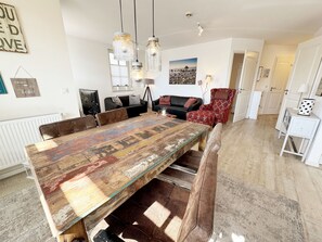 Ein geräumiger Essbereich mit einem großen Holztisch, mehreren Stühlen, Pendelleuchten und Blick auf das angrenzende Wohnzimmer.