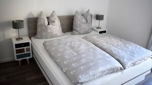 A double bed with grey bedding featuring anchor patterns, two bedside tables with lamps, and a small wooden nightstand with drawers. - 50.3.N Nordkoje - Modern, high-quality vacation apartment, sauna, large balcony, (Wangerland)