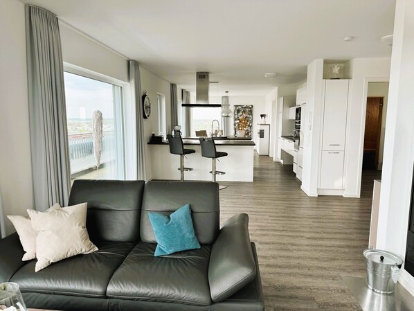 Un salon moderne avec un canapé en cuir noir, une cuisine en arrière-plan et une vue sur la ville grâce à de grandes fenêtres.