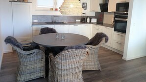 A modern kitchen with white cabinets, a round dining table with wicker chairs, and a unique hanging light fixture. - 49.4.M Ankerplatz - Modern, high-quality vacation apartment, sauna, large balcony, (Wangerland)