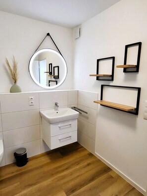 Un baño moderno con lavamanos blanco, espejo y suelo de madera. Hay tres estantes negros en la pared y un cubo de basura pequeño.