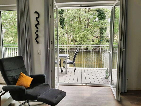 A room with a black chair and a yellow pillow, a sliding glass door leading to a balcony with a view of a river and trees. - 50.0.S Waterkant - High-quality vacation apartment, sauna, south-facing balcony. Graftlage, large (Wangerland)