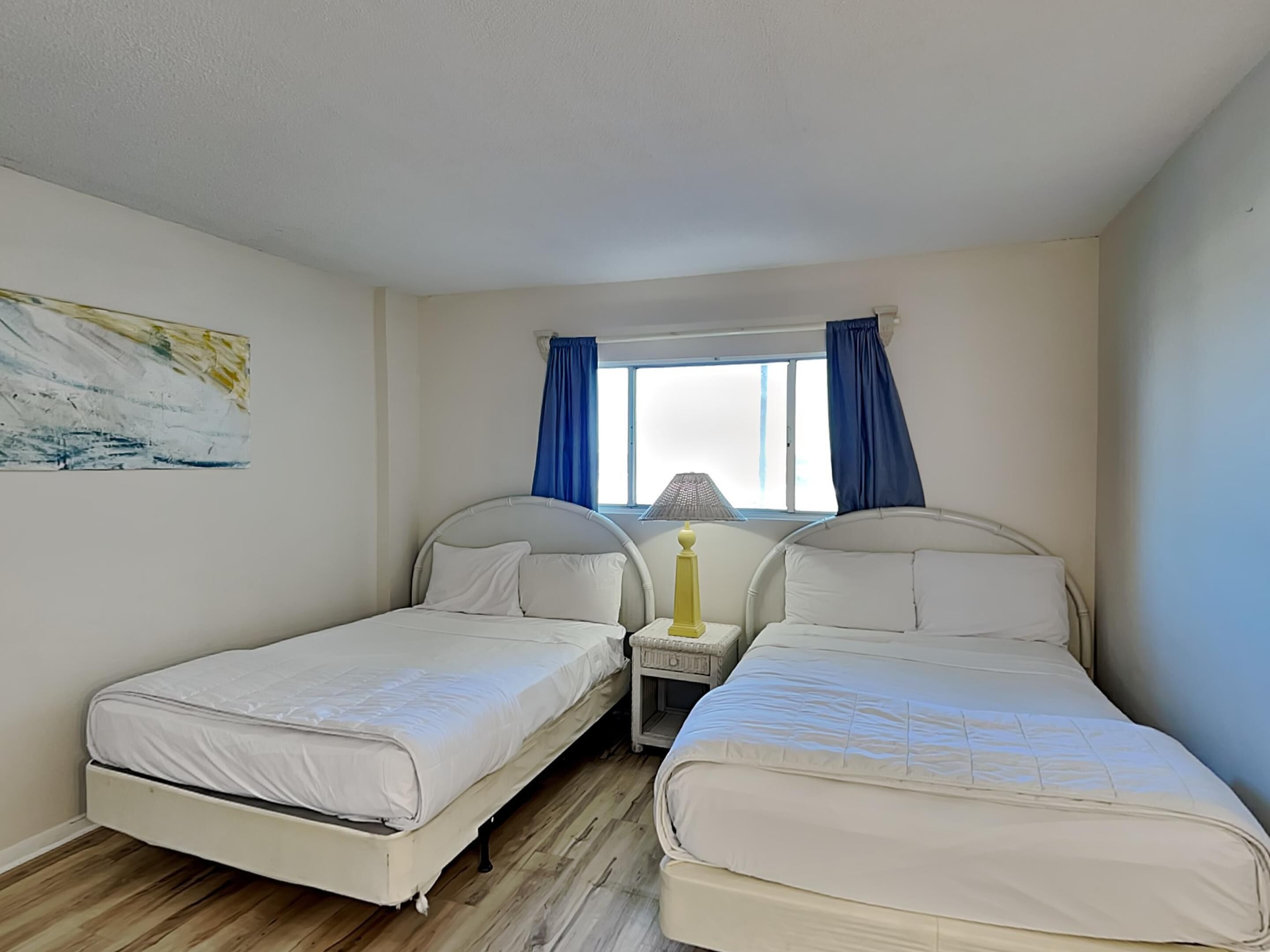 Une chambre avec deux lits simples, une petite table avec une lampe, une fenêtre avec des rideaux bleus et un tableau au mur.