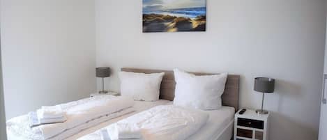 A double bed with white linens, two bedside tables with lamps, and a painting of a beach scene on the wall.