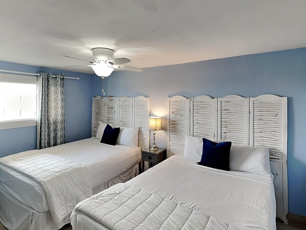 A hotel room with two beds, a ceiling fan, a window with curtains, and a nightstand with a lamp. - Regency Towers #803 by Resort Collection (Panama City Beach)