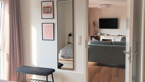 A modern hotel room with a large bed, a desk, a chair, a mirror, and a television in the adjacent living area.