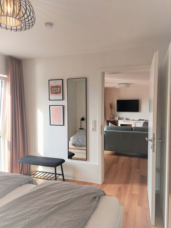 Et moderne hotelværelse med en stor seng, et skrivebord, en stol, et spejl og et fjernsyn i det tilstødende opholdsområde.