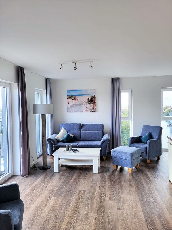 Un soggiorno moderno con un divano blu, un tavolino da caffè bianco e una poltrona blu. Appeso al muro, un quadro raffigura una spiaggia, e ampie finestre sono adornate da tende.