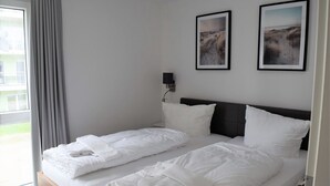Una cama doble con ropa blanca y almohadas, una mesita de noche con lámpara, y dos cuadros enmarcados en la pared.