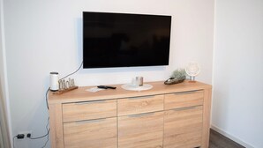 En moderne stue med et fladskærms-tv monteret på væggen, et træskab med skuffer, en lille plante og en pynteting på skabet.